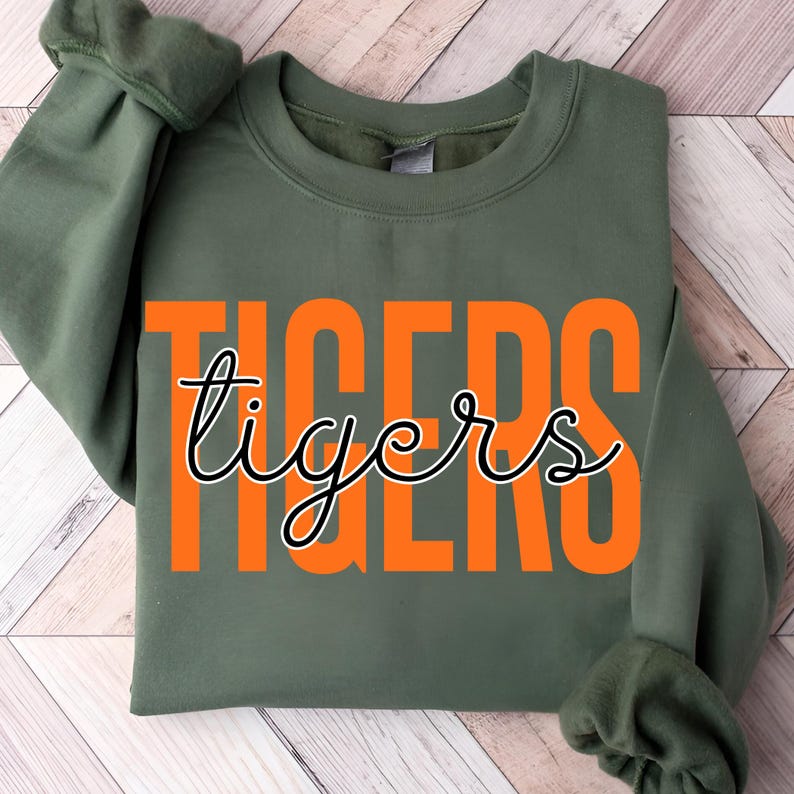 May include: An olive green sweatshirt with the word "TIGERS" in orange block letters. The word "tigers" is written in a cursive font over the orange letters. The sweatshirt has a crew neck and long sleeves.