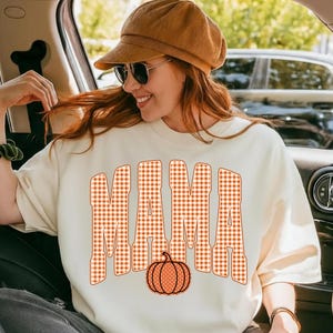 May include: A cream-colored short-sleeve t-shirt featuring the word MAMA in a large, orange and white gingham check pattern. Below the text is a graphic of a solid orange pumpkin with white polka dots. The shirt has a crew neckline.
