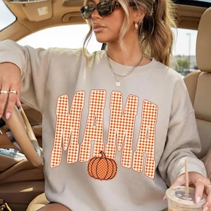 May include: A beige long-sleeve sweatshirt featuring the word MAMA printed in a large, orange and white gingham check pattern. Below the text is a graphic of an orange pumpkin with white polka dots. The sweatshirt has a crew neckline.