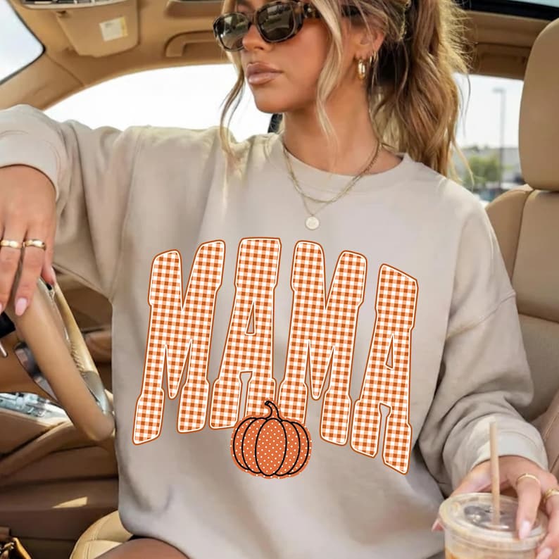 May include: A beige long-sleeve sweatshirt featuring the word MAMA printed in a large, orange and white gingham check pattern. Below the text is a graphic of an orange pumpkin with white polka dots. The sweatshirt has a crew neckline.