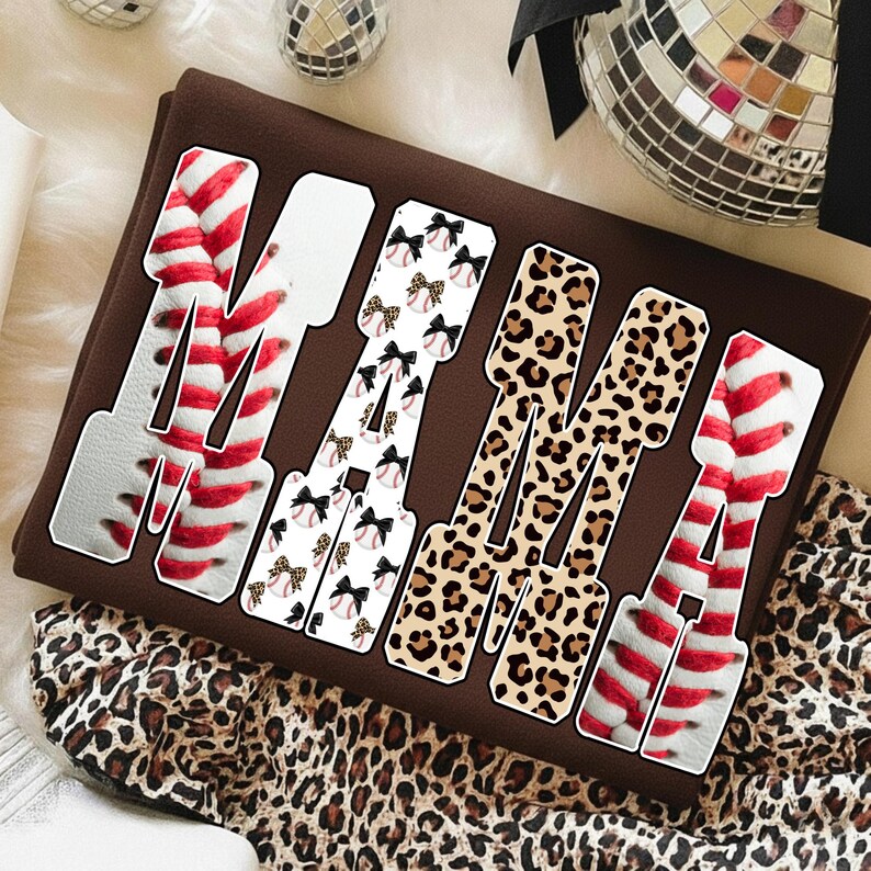 May include: A brown sweatshirt with the word "MAMA" in large, bold letters. The letters are decorated with baseball stitching, leopard print, and baseballs with bows. The sweatshirt is folded on a surface with a leopard print pattern.