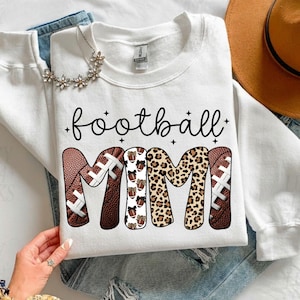 May include: White sweatshirt with the words "football mimi" in black script. The word "mimi" is designed with footballs and leopard print. A floral necklace and a brown hat are also visible.