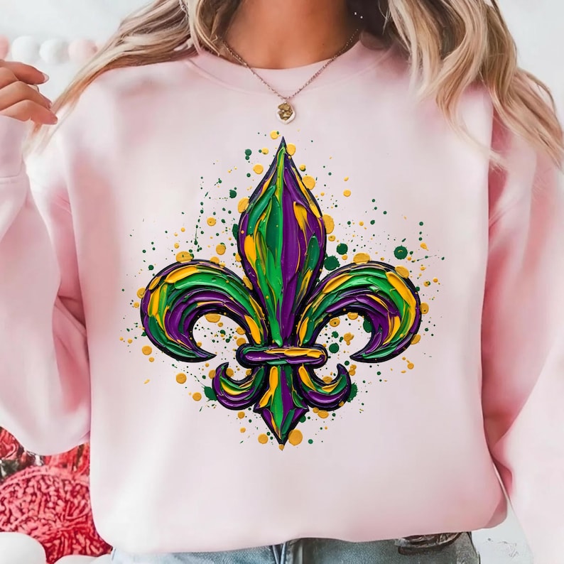 May include: A pale pink sweatshirt with a colourful fleur-de-lis emblem. The fleur-de-lis is painted in green, yellow, and purple, with a textured finish. Green and yellow paint splatters surround the design, creating a striking visual.