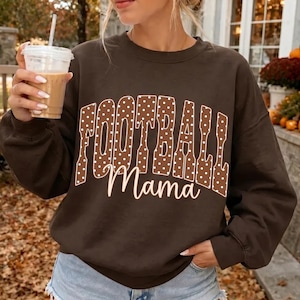May include: A dark brown long-sleeve sweatshirt featuring the word FOOTBALL in large, white, polka-dotted block letters, with the word Mama written underneath in a white cursive script font. The garment has a crew neckline and long sleeves.