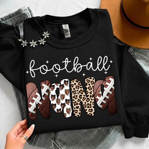 May include: Black sweatshirt with the words "football Nana" in white script. The word "Nana" is designed with footballs and leopard print. A necklace with flower accents is visible.