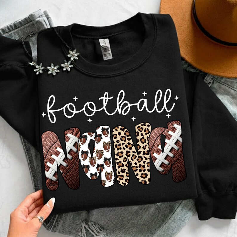 May include: Black sweatshirt with the words "football Nana" in white script. The word "Nana" is designed with footballs and leopard print. A necklace with flower accents is visible.
