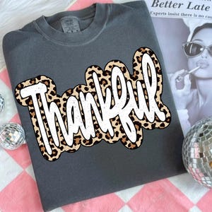 Peut inclure: Un sweat-shirt gris fonc&eacute; avec le mot "Thankful" en blanc, avec un motif l&eacute;opard sur le devant. Le sweat-shirt a un col rond et des manches longues. L'&eacute;tiquette de la marque est visible en haut.