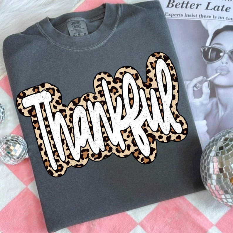 Peut inclure: Un sweat-shirt gris fonc&eacute; avec le mot "Thankful" en blanc, avec un motif l&eacute;opard sur le devant. Le sweat-shirt a un col rond et des manches longues. L'&eacute;tiquette de la marque est visible en haut.