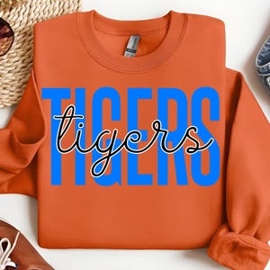 May include: An orange sweatshirt with the word "TIGERS" in large blue block letters, with the word "tigers" in a cursive font overlaying the blue letters. The sweatshirt is folded.