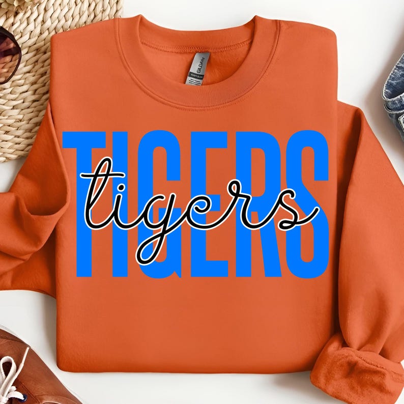 May include: An orange sweatshirt with the word "TIGERS" in large blue block letters, with the word "tigers" in a cursive font overlaying the blue letters. The sweatshirt is folded.