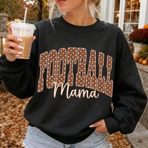 May include: A black long-sleeve sweatshirt featuring the word FOOTBALL in large brown letters with white polka dots, and the word Mama in white cursive script underneath. The garment has a crew neckline and long sleeves with ribbed cuffs.