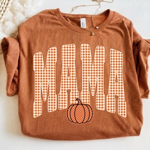 May include: A rust-colored short-sleeve t-shirt featuring the word MAMA printed in a white and orange gingham check pattern. Below the text is a graphic of a pumpkin with a white polka dot pattern on a rust-colored background with black outlines.