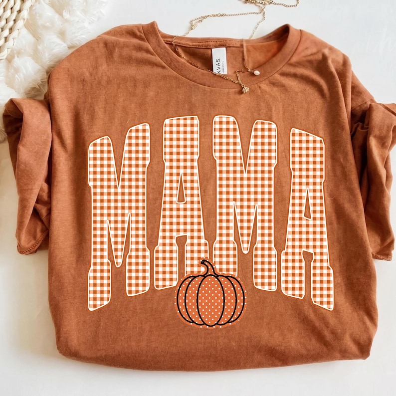 May include: A rust-colored short-sleeve t-shirt featuring the word MAMA printed in a white and orange gingham check pattern. Below the text is a graphic of a pumpkin with a white polka dot pattern on a rust-colored background with black outlines.