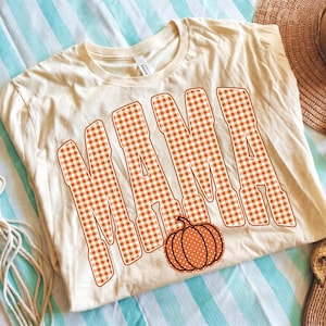May include: A cream-colored short-sleeve t-shirt featuring the word MAMA printed in large, bold letters with an orange and white gingham check pattern. Below the text, there is a small, round orange pumpkin graphic with white polka dots.