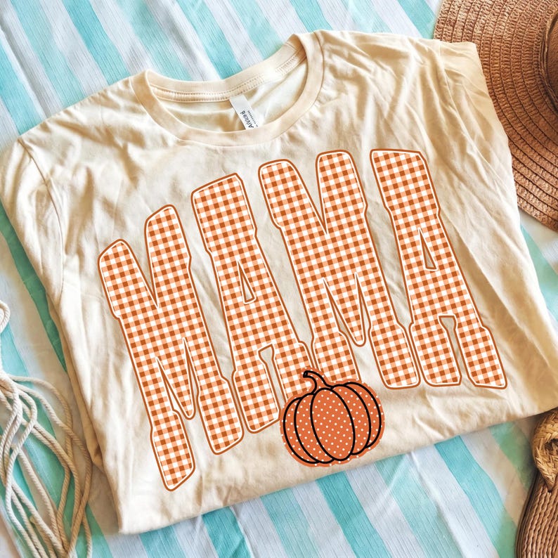 May include: A cream-colored short-sleeve t-shirt featuring the word MAMA printed in large, bold letters with an orange and white gingham check pattern. Below the text, there is a small, round orange pumpkin graphic with white polka dots.