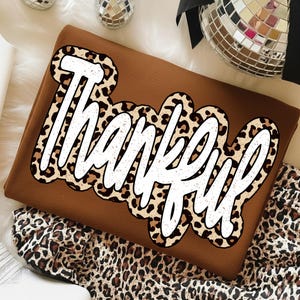 Op de afbeelding: Een bruine sweatshirt met het woord "Thankful" in een witte, vette letter met een luipaardprint rand. De sweatshirt is op een wit oppervlak gevouwen, met luipaardprint stof zichtbaar.