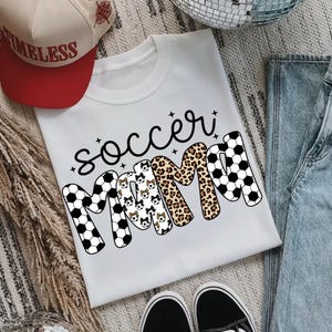May include: White t-shirt with the words "Soccer Mama" in black script. The word "MAMA" is designed with soccer ball patterns, leopard print, and dog illustrations. A red and tan baseball cap with the word "TIMELESS" is visible.