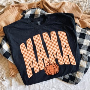 May include: A black short-sleeved t-shirt featuring the word MAMA in large, orange and white gingham-patterned block letters. Below the text is a small, orange pumpkin graphic with white polka dots, centered on the front of the shirt.