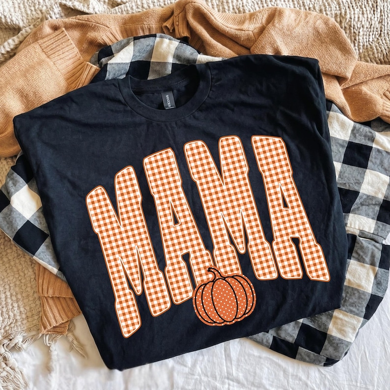 May include: A black short-sleeved t-shirt featuring the word MAMA in large, orange and white gingham-patterned block letters. Below the text is a small, orange pumpkin graphic with white polka dots, centered on the front of the shirt.