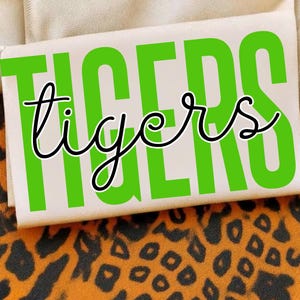 May include: A rectangular white object with the word "TIGERS" in large, bright green capital letters and the word "tigers" in a cursive font. The object is set against a background of tan fabric and a leopard print.