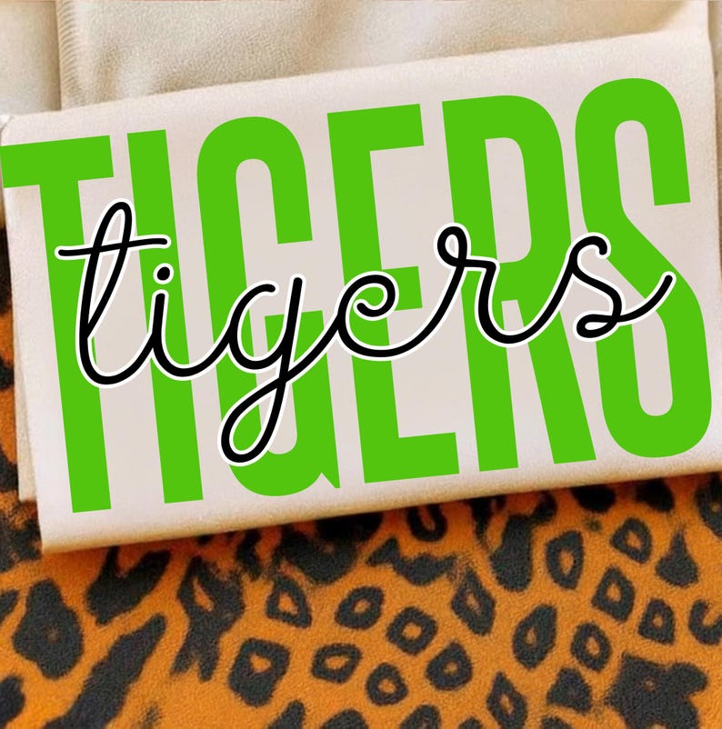 May include: A rectangular white object with the word "TIGERS" in large, bright green capital letters and the word "tigers" in a cursive font. The object is set against a background of tan fabric and a leopard print.