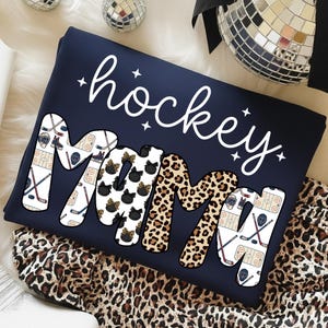May include: Navy blue sweatshirt with the words "hockey mama" in white script. The word "mama" is decorated with hockey-themed graphics, black bows, and leopard print. The sweatshirt is folded on a leopard print fabric.