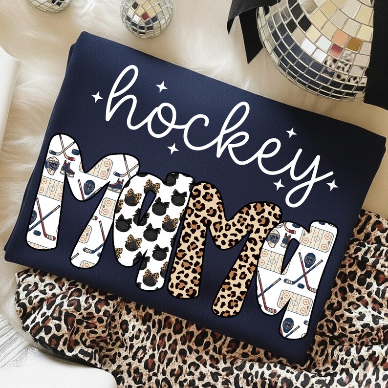 May include: Navy blue sweatshirt with the words "hockey mama" in white script. The word "mama" is decorated with hockey-themed graphics, black bows, and leopard print. The sweatshirt is folded on a leopard print fabric.