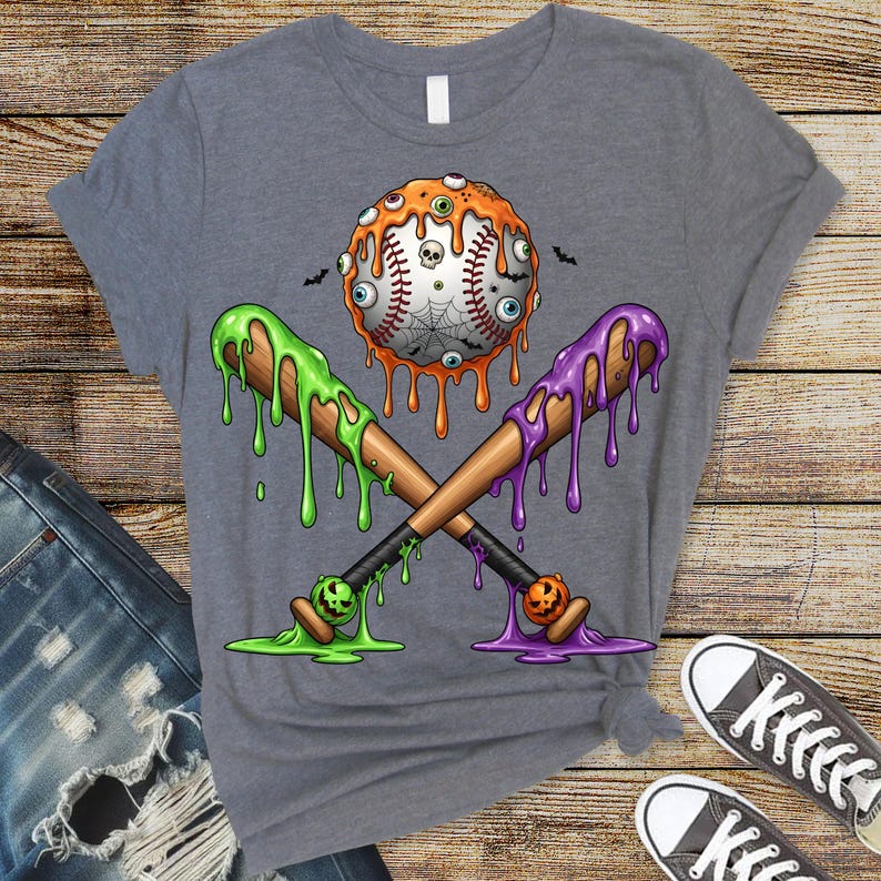 K&ouml;nnte beinhalten: Graues T-Shirt mit einem Halloween-Motiv. Das Design zeigt einen Baseball mit Aug&auml;pfeln, einem Spinnennetz und einem Totenkopf, gekreuzte Baseballschl&auml;ger mit gr&uuml;nem und lila Schleim und kleine Flederm&auml;use.