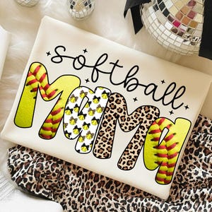 May include: Cream-colored t-shirt with the words "Softball Mom" in black script. The word "Mom" is designed with softball and leopard print patterns. The shirt is folded on a leopard print fabric.
