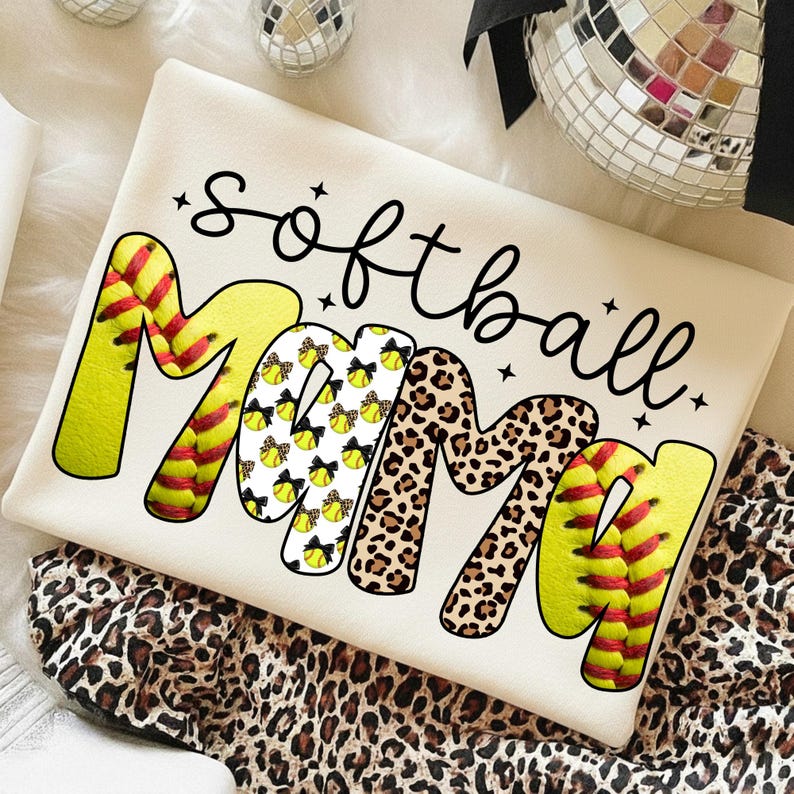 May include: Cream-colored t-shirt with the words "Softball Mom" in black script. The word "Mom" is designed with softball and leopard print patterns. The shirt is folded on a leopard print fabric.