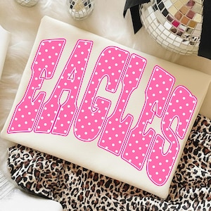 Pode incluir: Uma camisola de cor creme com a palavra EAGLES em letras grandes, arrojadas e cor-de-rosa com um padrão de bolinhas brancas. As letras têm um contorno rosa espesso e um design de fonte ondulado. A peça de vestuário está dobrada e plana.