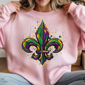 May include: A pale pink sweatshirt displaying a colourful fleur-de-lis emblem. The design uses green, purple, and yellow hues, with paint splatter effects. The sweatshirt is a casual item of clothing.