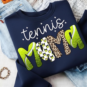 K&ouml;nnte beinhalten: Marineblaues Sweatshirt mit dem Schriftzug "tennis mama" in wei&szlig;er Schrift. Das Wort "mama" zeigt Tennisball- und Leopardenmuster. Das Sweatshirt ist auf einer blauen Jeans und einer cremefarbenen Decke gefaltet.