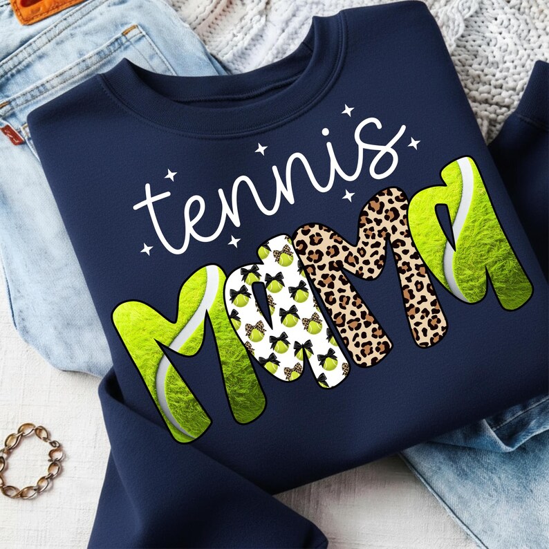 K&ouml;nnte beinhalten: Marineblaues Sweatshirt mit dem Schriftzug "tennis mama" in wei&szlig;er Schrift. Das Wort "mama" zeigt Tennisball- und Leopardenmuster. Das Sweatshirt ist auf einer blauen Jeans und einer cremefarbenen Decke gefaltet.