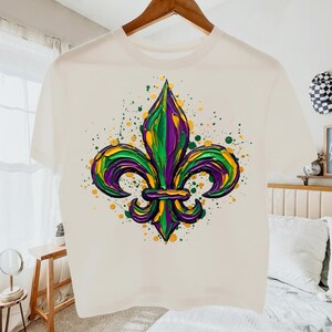 May include: A white t-shirt with a colourful fleur-de-lis design in green, purple, and yellow, with paint splatter details. The shirt is displayed on a wooden hanger.