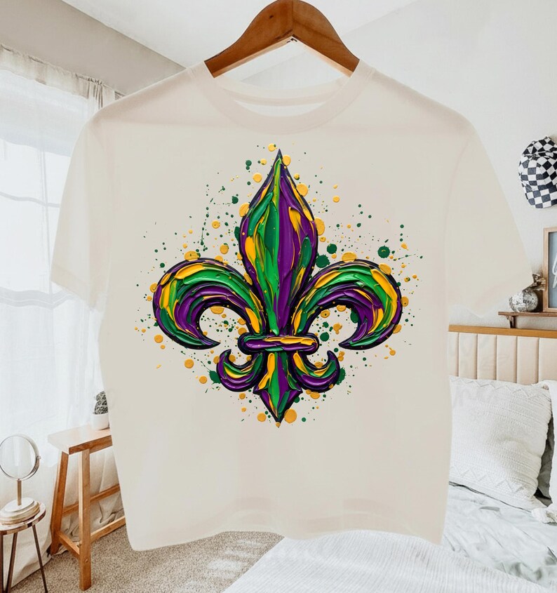 May include: A white t-shirt with a colourful fleur-de-lis design in green, purple, and yellow, with paint splatter details. The shirt is displayed on a wooden hanger.