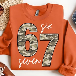 May include: An orange sweatshirt with the numbers "67" in a camouflage pattern. The word "six" is written above the number "6" and the word "seven" is written below the number "7", both in white script.