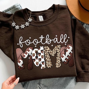May include: A brown sweatshirt with the words "football mama" in white script. The word "mama" is designed with footballs and leopard print. A silver necklace with flower accents is visible.