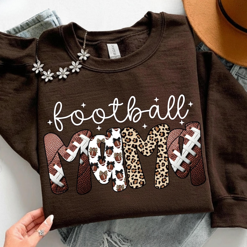 May include: A brown sweatshirt with the words "football mama" in white script. The word "mama" is designed with footballs and leopard print. A silver necklace with flower accents is visible.
