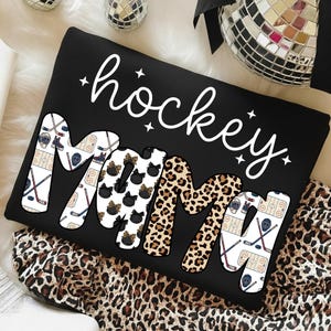 May include: Black sweatshirt with the words "hockey mama" in white script and patterned letters. The "M"s feature hockey pucks and leopard print. Hockey sticks and ice rink graphics decorate the other letters.