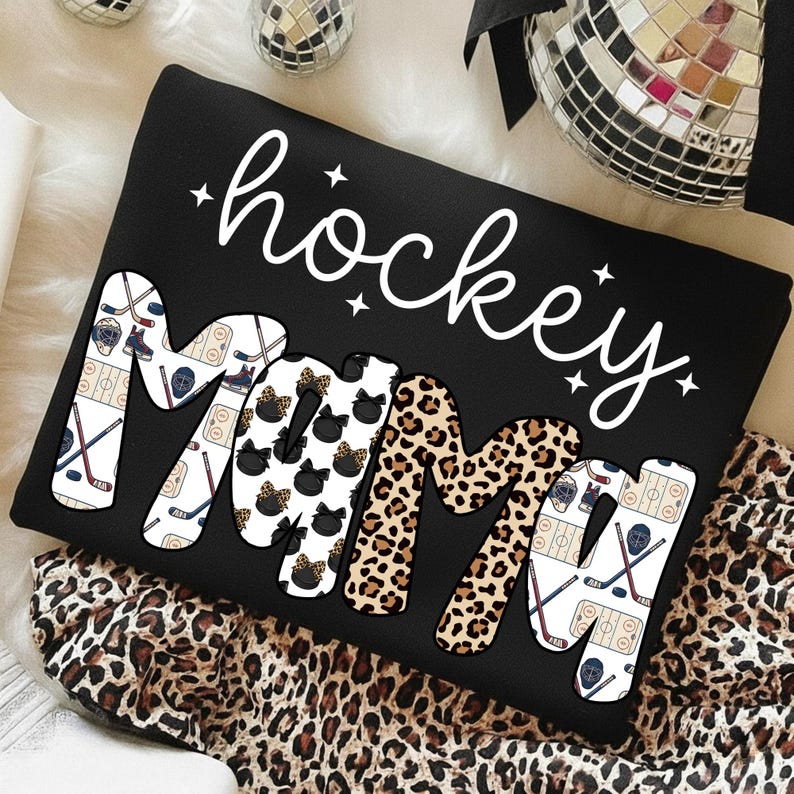 May include: Black sweatshirt with the words "hockey mama" in white script and patterned letters. The "M"s feature hockey pucks and leopard print. Hockey sticks and ice rink graphics decorate the other letters.