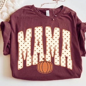May include: A maroon short-sleeved t-shirt featuring the word MAMA in large, cream-colored block letters with a brown polka dot pattern. Below the text is a graphic of a pumpkin with the same cream and brown polka dot design.