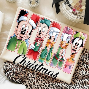 Puede incluir: Camiseta color crema con la palabra "MERRY" y los personajes de Disney Mickey, Minnie, Donald, Daisy y Goofy, todos con atuendo navideño. La palabra "Christmas" está escrita debajo en cursiva.