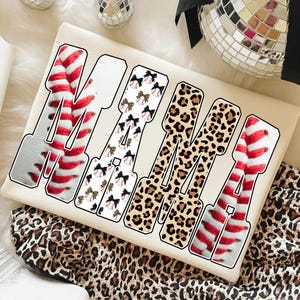 May include: Cream-colored t-shirt with the word "MAMA" in a bold, outlined font. The letters are filled with various patterns: baseball stitching, baseballs with bows, and leopard print. The shirt is on a leopard print fabric.