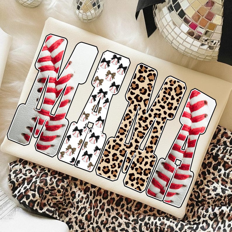 May include: Cream-colored t-shirt with the word "MAMA" in a bold, outlined font. The letters are filled with various patterns: baseball stitching, baseballs with bows, and leopard print. The shirt is on a leopard print fabric.