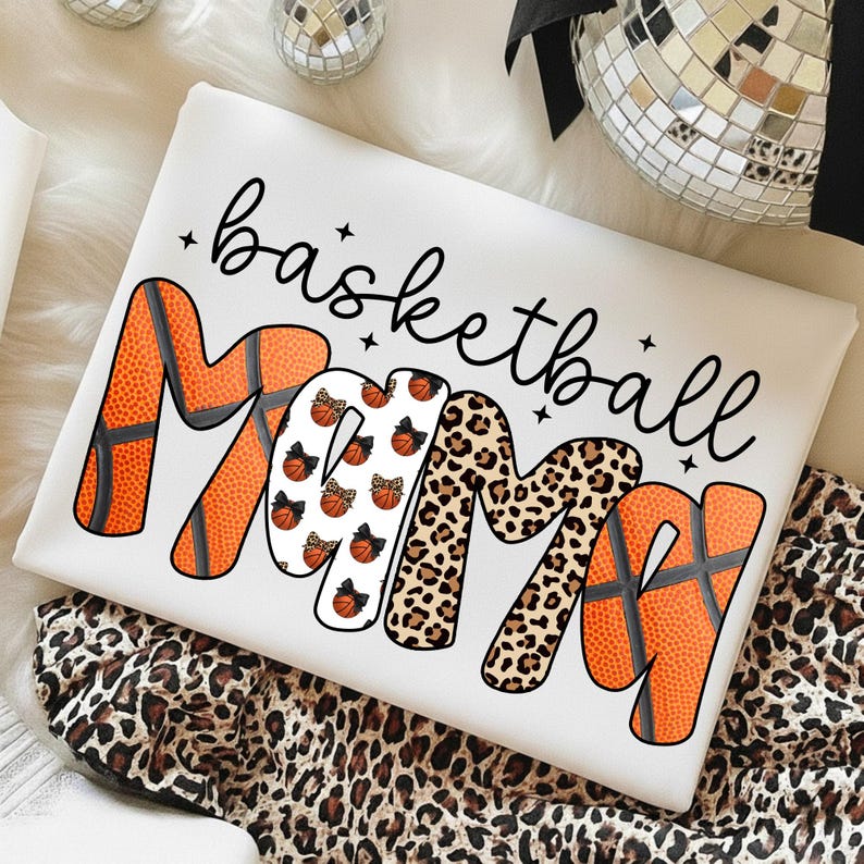 May include: White t-shirt with the words "basketball mama" in black script. The word "MAMA" is designed with basketball textures, leopard print, and basketball graphics. The shirt is displayed on a leopard print fabric.