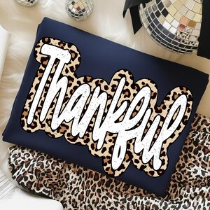 Peut inclure: Sweat-shirt bleu marine avec le mot "Thankful" en &eacute;criture blanche, bord&eacute; d'un motif l&eacute;opard. Le sweat-shirt est pli&eacute; sur une surface blanche, avec un tissu &agrave; imprim&eacute; l&eacute;opard visible.