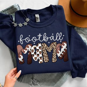 May include: Navy blue sweatshirt with the words "football mama" in white script. The word "MAMA" is designed with footballs and leopard print. A necklace with flower accents and a brown hat are also visible.