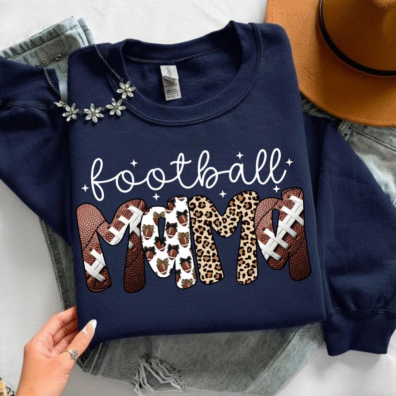 May include: Navy blue sweatshirt with the words "football mama" in white script. The word "MAMA" is designed with footballs and leopard print. A necklace with flower accents and a brown hat are also visible.