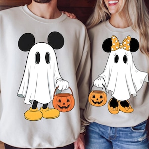 May include: Beige sweatshirts featuring a cartoon ghost design. The ghost is wearing Mickey Mouse ears and holding a jack-o'-lantern bucket. One ghost has a bow. The sweatshirts are perfect for Halloween.